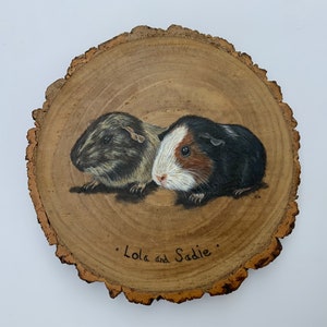 May include: A wood slice with a painted image of two guinea pigs. The guinea pigs are brown, black, and white. The wood slice has the text "Lola and Sadie" written on it.