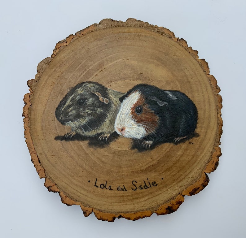 May include: A wood slice with a painted image of two guinea pigs. The guinea pigs are brown, black, and white. The wood slice has the text "Lola and Sadie" written on it.