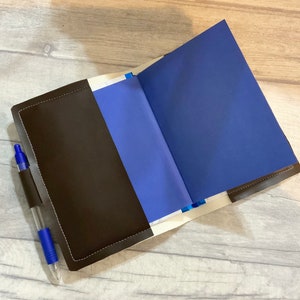 Peut inclure: Un carnet en cuir marron avec un int&eacute;rieur bleu et un stylo bleu qui sort de la poche lat&eacute;rale.