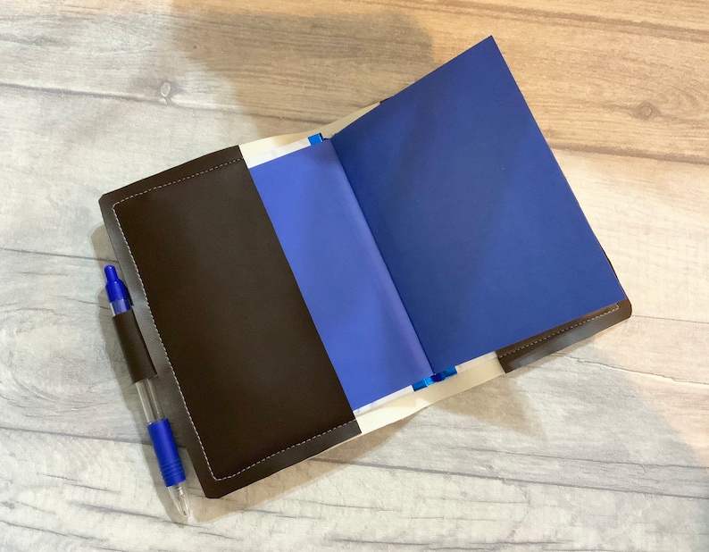 Peut inclure: Un carnet en cuir marron avec un int&eacute;rieur bleu et un stylo bleu qui sort de la poche lat&eacute;rale.