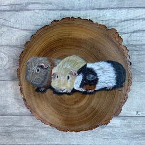 May include: A painting of three guinea pigs on a wooden slice. The guinea pigs are brown, white, and black with brown eyes. The wooden slice is brown with a natural wood grain.