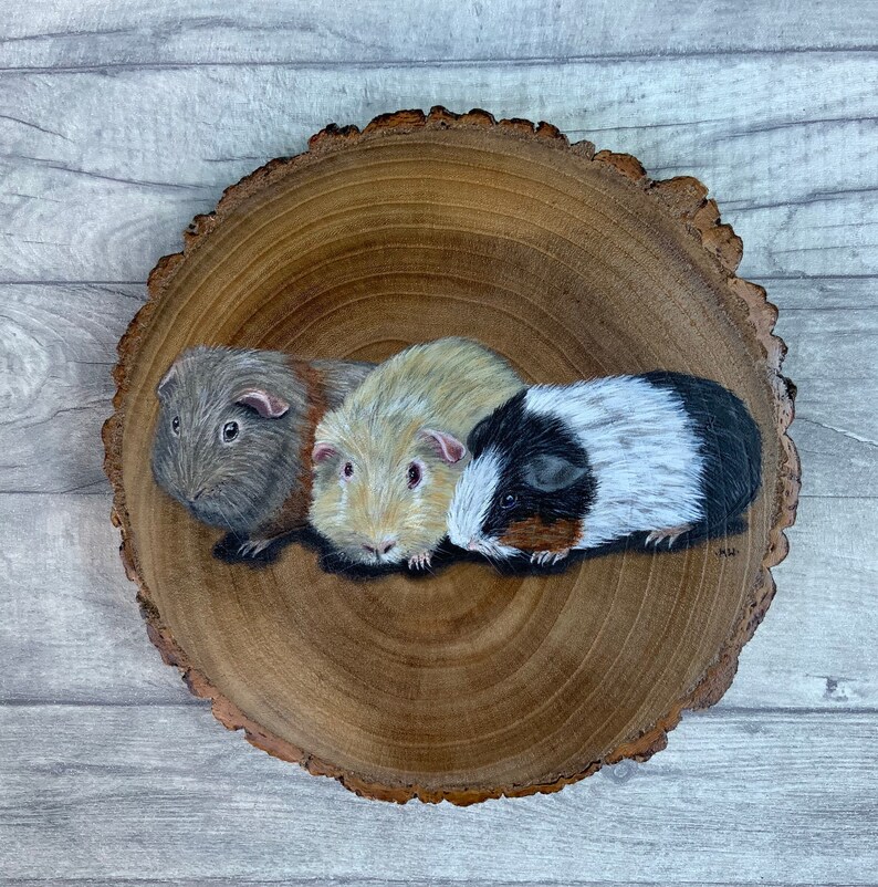 May include: A painting of three guinea pigs on a wooden slice. The guinea pigs are brown, white, and black with brown eyes. The wooden slice is brown with a natural wood grain.