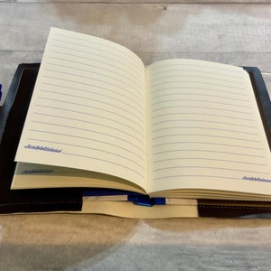 Peut inclure: Un carnet reli&eacute; en cuir noir avec un porte-stylo et des pages lign&eacute;es. Le carnet est ouvert sur une page blanche avec le mot "Scribblelicious" &eacute;crit &agrave; l'encre bleue.