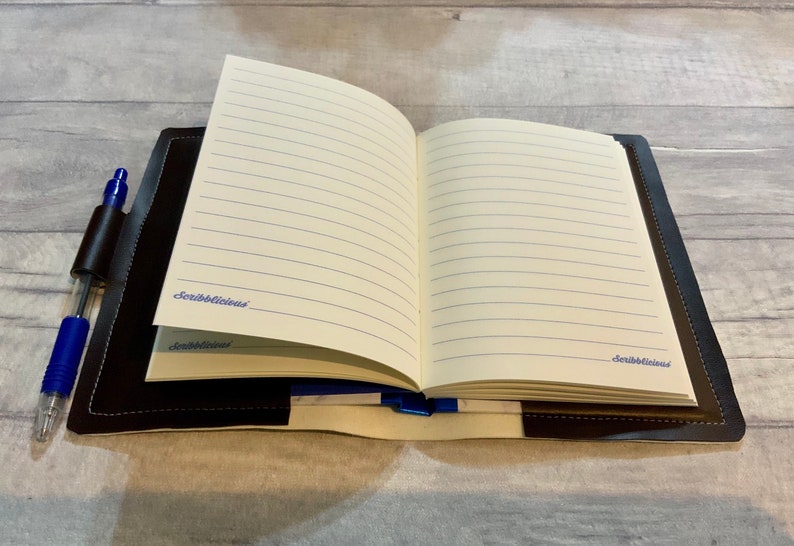 Peut inclure: Un carnet reli&eacute; en cuir noir avec un porte-stylo et des pages lign&eacute;es. Le carnet est ouvert sur une page blanche avec le mot "Scribblelicious" &eacute;crit &agrave; l'encre bleue.