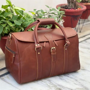 May include: A brown leather duffel bag featuring two top handles with a central leather grip, a fold-over flap with a buckle closure, brass-toned metal hardware, and a side zipper pocket. The bag has vertical leather straps and reinforced stitching.