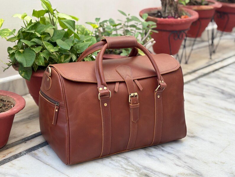 May include: A brown leather duffel bag featuring two top handles with a central leather grip, a fold-over flap with a buckle closure, brass-toned metal hardware, and a side zipper pocket. The bag has vertical leather straps and reinforced stitching.