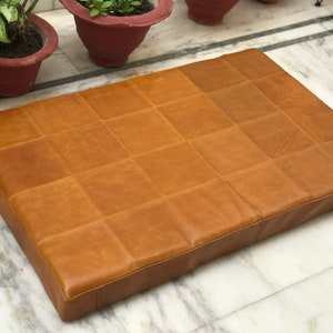May include: A large, rectangular, tan leather cushion with a quilted design. The cushion is made of multiple squares of leather sewn together.