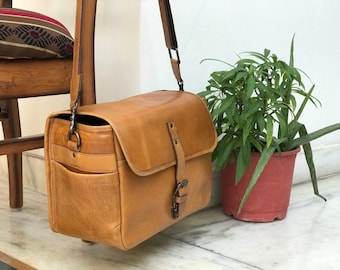 May include: A tan leather messenger bag with a long strap and a buckle closure. The bag has a front pocket and a flap closure.