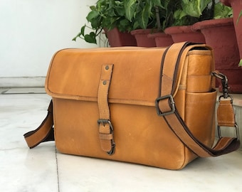 May include: A tan leather camera bag with a long adjustable strap and a buckle closure. The bag has a flap with a buckle closure and a small pocket on the front.