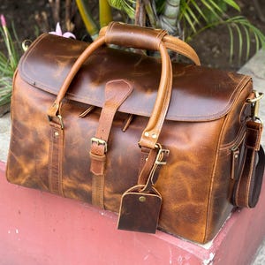 May include: A brown leather duffel bag featuring a large main compartment with a flap closure, two top handles, and a central buckle strap. The bag includes a detachable shoulder strap, brass-toned hardware, and a matching leather luggage tag.
