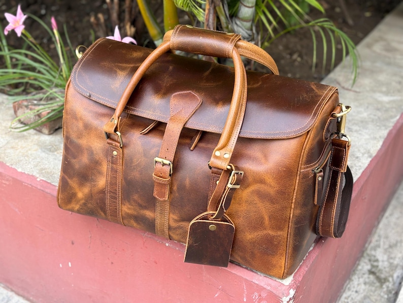 May include: A brown leather duffel bag featuring a large main compartment with a flap closure, two top handles, and a central buckle strap. The bag includes a detachable shoulder strap, brass-toned hardware, and a matching leather luggage tag.
