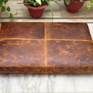 May include: A brown leather floor cushion with a square shape and a stitched design. The cushion is made of distressed leather and has a rustic look.