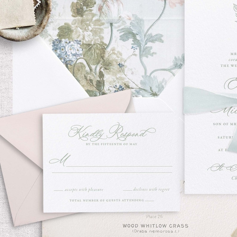 May include: A white RSVP card with a pale pink envelope. The card features green script writing that reads "Kindly Respond by the Fifteenth of May". There are lines for the guest to fill in their name, whether they accept or decline the invitation, and the total number of guests attending.