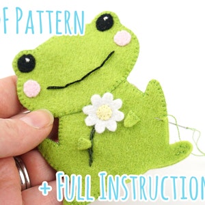 Könnte beinhalten: Ein grüner Filz-Frosch, der eine weiße Filz-Blume hält. Der Frosch hat schwarze Augen, eine rosa Wange und ein schwarzes gesticktes Lächeln. Der Text "PDF PATTERN + FULL INSTRUCTIONS" ist unten im Bild sichtbar.