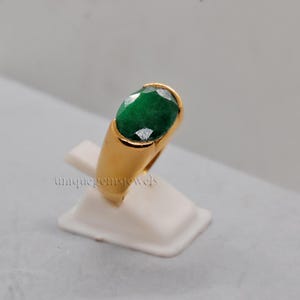 Könnte beinhalten: Ein goldener Ring mit einem großen, ovalen, grünen Edelstein. Der Ring befindet sich auf einem weißen Präsentationsständer.