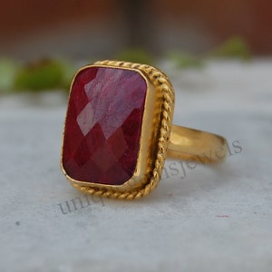 Puede incluir: Un anillo de oro con una gran piedra preciosa roja rectangular. El anillo tiene un diseño de cuerda alrededor de la banda.