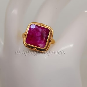 May include: A gold ring featuring a square-shaped ruby gemstone. The ring has a textured band and a square setting.