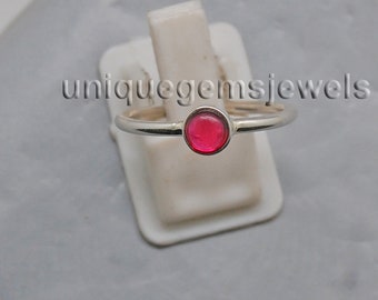 Könnte beinhalten: Ein silberner Ring mit einem runden, leuchtend roten Edelstein. Der Ring wird auf einem weißen Ständer vor grauem Hintergrund präsentiert. Der Text "uniquegemsjewels" ist sichtbar.