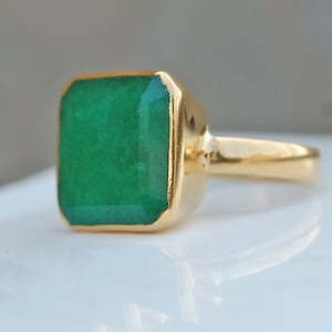 Könnte beinhalten: Ein goldener Ring mit einem großen, smaragdgrünen Edelstein. Der Edelstein ist in einer quadratischen Form gefasst und hat eine polierte Oberfläche.
