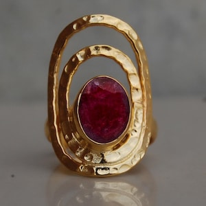 May include: A gold-colored ring with a large, oval-shaped, deep red gemstone at the center. The ring has a hammered texture and a unique design with two concentric oval shapes surrounding the gemstone. The ring is a piece of jewelry.
