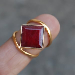 May include: A gold ring with a red gemstone set in a square shape. The ring has a simple design with a single band.