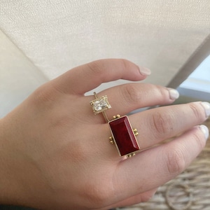 May include: Two gold rings with gemstones. The ring on the left has a clear rectangular stone. The ring on the right has a dark red rectangular stone.