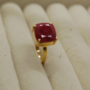 Pode incluir: Um anel de ouro com uma grande gema vermelha de forma quadrada.