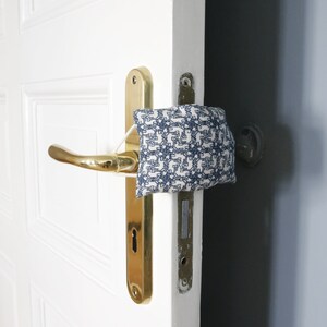 May include: A white door with a gold door handle and a blue and white patterned fabric doorstop. The doorstop is placed on the handle to prevent the door from opening.
