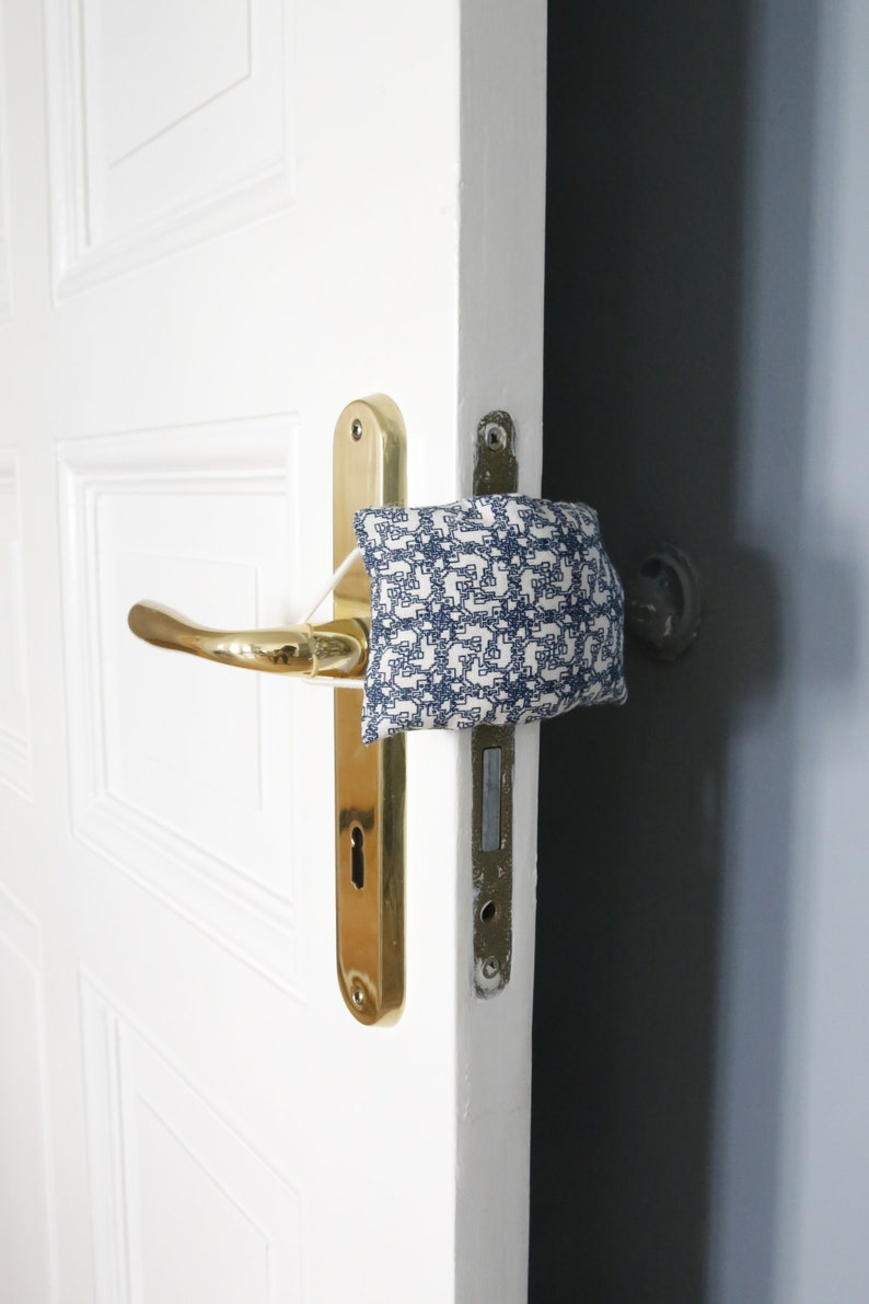 May include: A white door with a gold door handle and a blue and white patterned fabric doorstop. The doorstop is placed on the handle to prevent the door from opening.