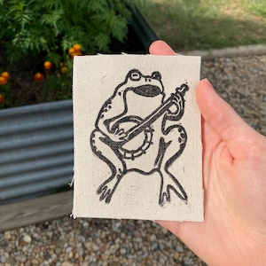 Könnte beinhalten: Ein quadratischer, cremefarbener Stoffaufnäher mit einer schwarzen Illustration einer Frosch, der ein Banjo spielt. Der Frosch ist sitzend dargestellt, mit gespreizten Beinen und Armen, die das Instrument halten. Der Aufnäher wird von einer Hand gehalten.