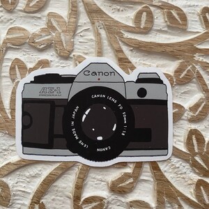 May include: A vintage Canon AE-1 Program camera sticker with a black and white illustration of the camera. The sticker features the text "Canon Lens FD 50mm 1:1.8" and "Lens Made in Japan".