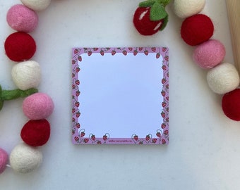 May include: A pink notepad with a white interior and a border of small red strawberries. The notepad has the text "mila-seventh co." printed on the bottom.