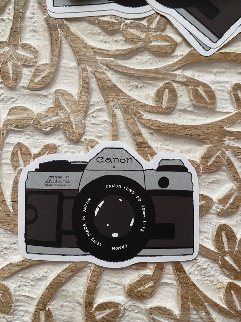 May include: A vintage Canon AE-1 Program camera sticker with a black and white illustration of the camera. The sticker features the text "Canon Lens FD 50mm 1:1.8" and "Lens Made in Japan".