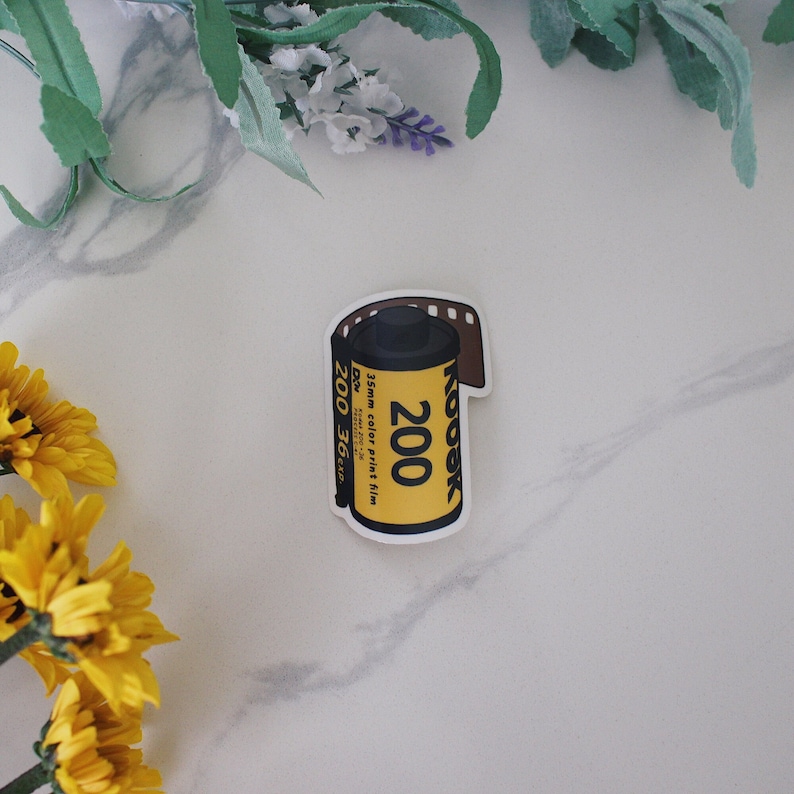 May include: A yellow Kodak 200 35mm color print film sticker. The sticker has a black top and a brown film strip. The text on the sticker reads "Kodak 200" and "35mm color print film".