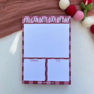 May include: A pink and white checkered notepad with the text "YOU CAN DO IT. DAY BY DAY." at the top. The notepad is divided into three sections: a large section at the top and two smaller sections at the bottom labeled "REMINDERS" and "TO DO". The notepad is on a white surface.