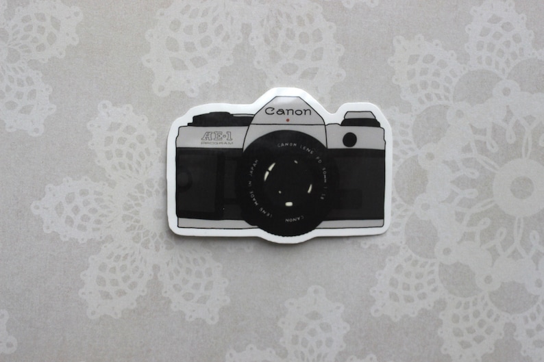 May include: A black and white sticker of a Canon AE-1 camera with a silver lens. The sticker has a white border.