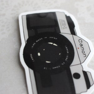 May include: A black and white sticker of a Canon AE-1 camera. The sticker features the Canon logo and the text "Canon Lens Made in Japan".