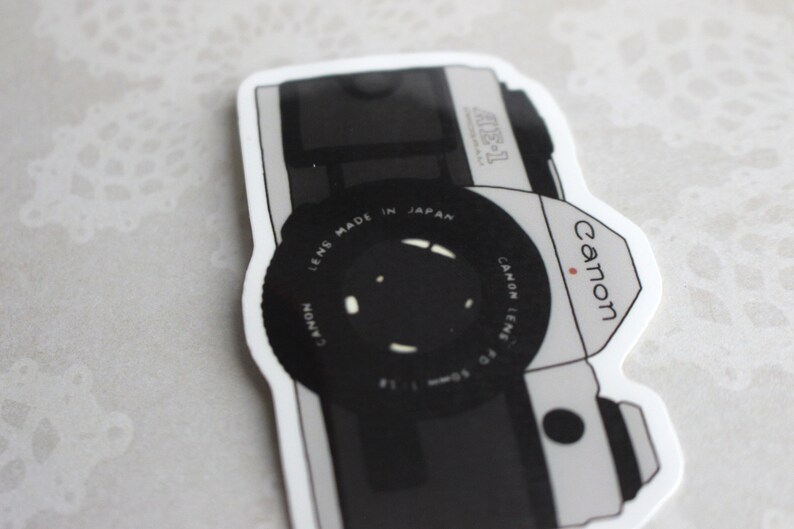 May include: A black and white sticker of a Canon AE-1 camera. The sticker features the Canon logo and the text "Canon Lens Made in Japan".