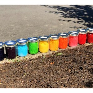 May include: Ten glass jars with lids, lined up in a row on a brown surface. The jars are filled with different colored substances, creating a rainbow effect. The colors are, from left to right, dark blue, purple, light blue, green, yellow, orange, red, pink, dark red, and brown.