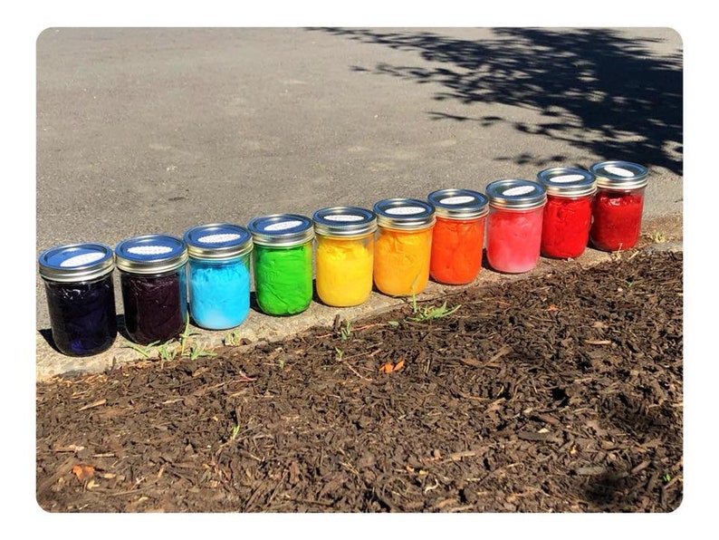 May include: Ten glass jars with lids, lined up in a row on a brown surface. The jars are filled with different colored substances, creating a rainbow effect. The colors are, from left to right, dark blue, purple, light blue, green, yellow, orange, red, pink, dark red, and brown.