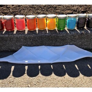May include: A row of 10 glass jars with lids, filled with different colored liquids, arranged in a rainbow pattern. The jars are sitting on a concrete curb with a white cloth lying in front of them.