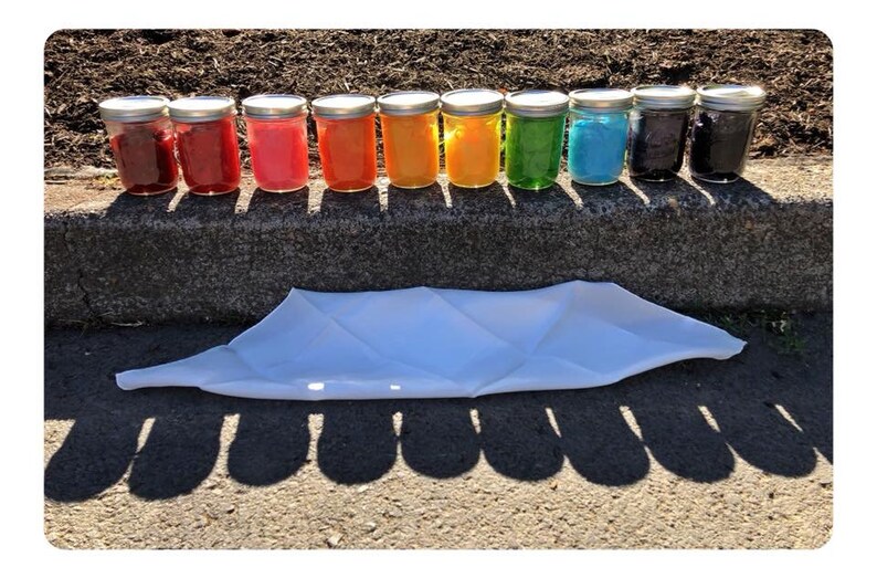 May include: A row of 10 glass jars with lids, filled with different colored liquids, arranged in a rainbow pattern. The jars are sitting on a concrete curb with a white cloth lying in front of them.