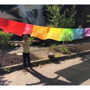 May include: A child stands in front of a rainbow-colored fabric banner hanging on a clothesline. The banner is made of several rectangular pieces of fabric, each a different color of the rainbow. The child is wearing a yellow shirt and blue pants and has their arms outstretched.