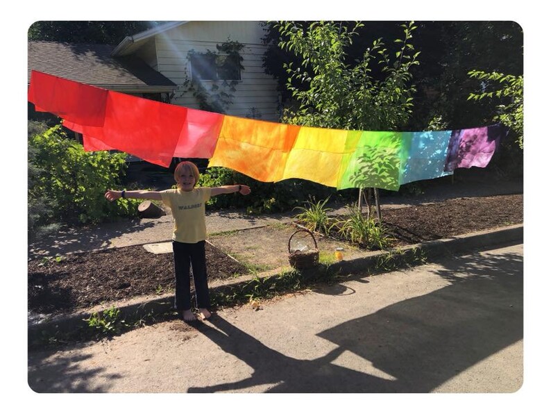 May include: A child stands in front of a rainbow-colored fabric banner hanging on a clothesline. The banner is made of several rectangular pieces of fabric, each a different color of the rainbow. The child is wearing a yellow shirt and blue pants and has their arms outstretched.