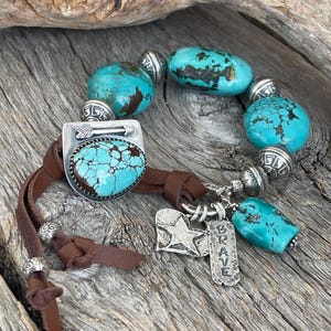 May include: A turquoise and silver bracelet with a leather cord and charms. The charms include a star, an arrow, and a tag that says "BRAVE".
