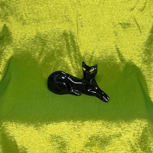 May include: A black ceramic cat figurine with bright yellow eyes, in a relaxed, reclining pose. The cat is glossy and rests on bright green, textured fabric. The figurine is a decorative item.