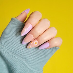 Pode incluir: Close-up de uma mão com unhas de cor lilás claro e nude com um design de flor de margarida. As unhas são longas e de forma oval.