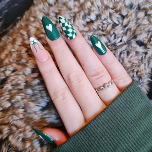Pode incluir: Close-up de uma mão com desenhos de esmalte verde e branco. As unhas são longas e pontiagudas, e os desenhos incluem corações, padrões xadrez e laços. A mão está usando um anel de prata com pequenas pedras redondas.