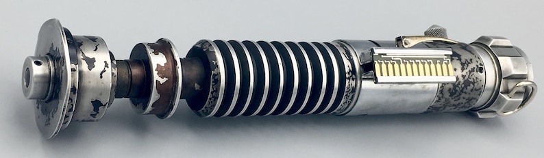May include: A silver lightsaber prop with a black and silver handle and a silver blade. The lightsaber has a weathered, distressed look.