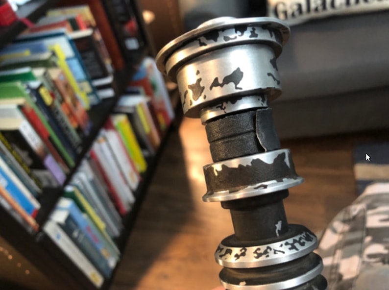 K&ouml;nnte beinhalten: Eine Nahaufnahme einer silbernen und schwarzen Lichtschwert-Griff mit einer verwitterten, abgenutzten Oberfl&auml;che. Der Griff hat eine Reihe von Ringen und einen schwarzen Griff. Der Griff ist mit schwarzer und silberner Farbe bedeckt, was ihm ein abgenutztes und gebrauchtes Aussehen verleiht.